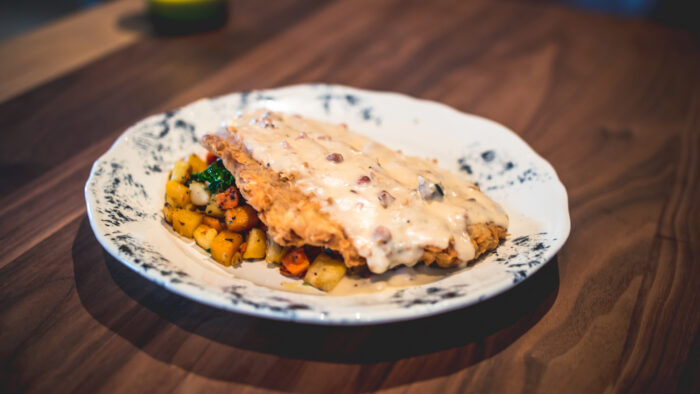 A plate of chicken fried steak topped with creamy gravy, served on a bed of sautéed diced vegetables including potatoes and carrots, on a white patterned plate over a wooden table.
