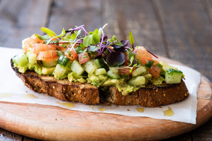 A slice of multigrain toast topped with mashed avocado, diced cucumber, tomato, and microgreens, served on parchment paper atop a wooden board.