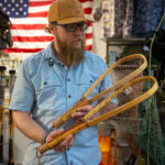 A bearded man wearing a tan cap and glasses examines wooden fishing nets in a store, with an American flag and fishing gear visible in the background.