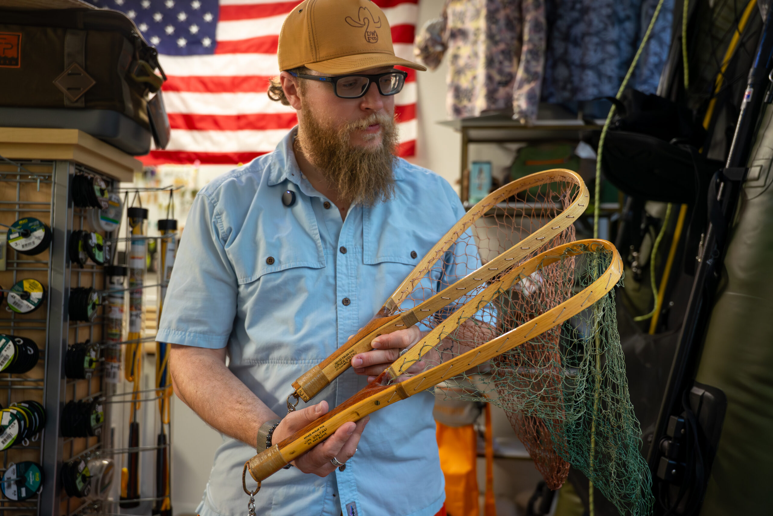 A bearded man wearing a tan cap and glasses examines wooden fishing nets in a store, with an American flag and fishing gear visible in the background.