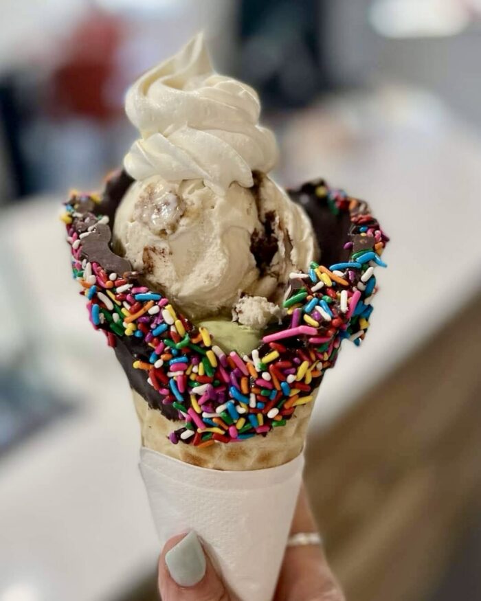 A hand holds an ice cream cone with chocolate-dipped edges covered in colorful sprinkles, two scoops of delicious ice cream, and a swirl of soft serve on top, all wrapped in a white napkin.