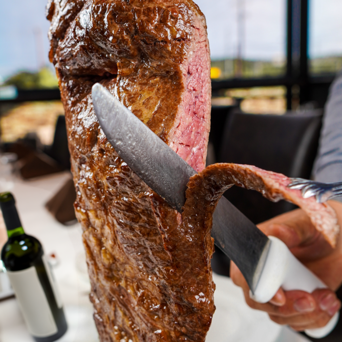 A close-up of a person slicing a piece of juicy, medium-rare grilled meat with a knife and fork at a restaurant table; a bottle of wine is visible in the background.