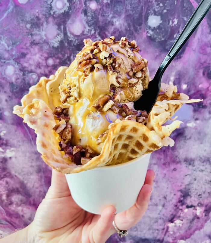 A hand holds a cup with a waffle bowl filled with two scoops of ice cream, topped with caramel sauce and chopped pecans, against a purple marbled background. A black spoon is tucked into the creamy ice cream delight.