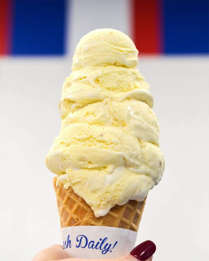 A hand holding a waffle cone with five scoops of vanilla ice cream, slightly melting, against a white background with blue and red stripes at the top.