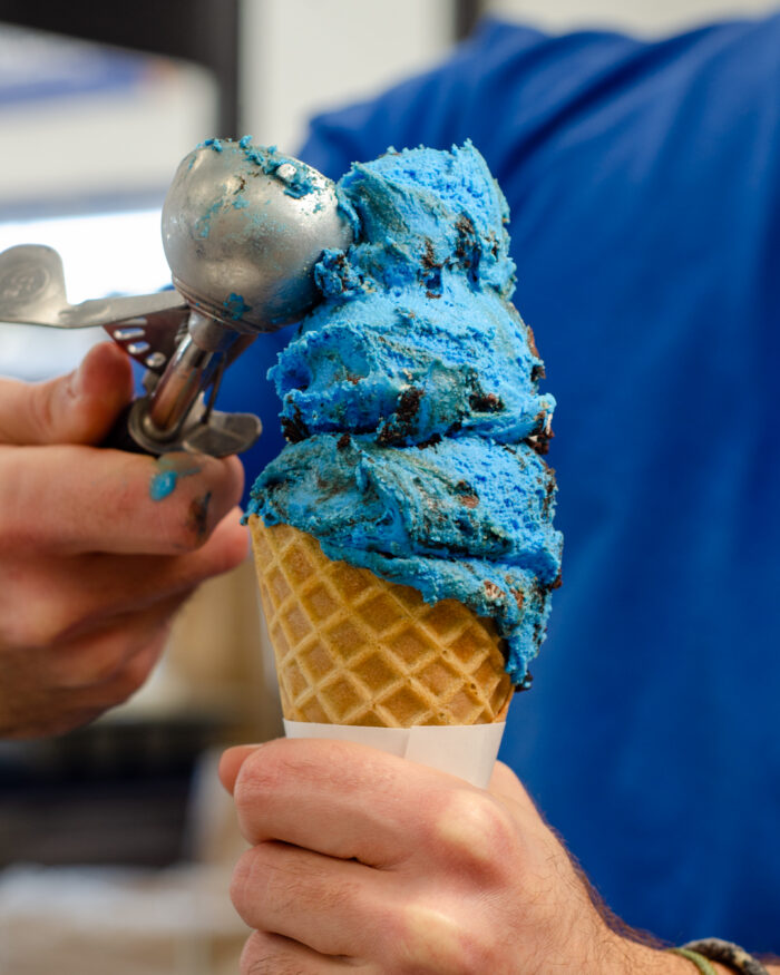 A person scoops bright blue ice cream with chocolate chunks onto a waffle cone, holding both the cone and the metal scoop. The person is wearing a blue shirt.