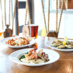 Three plates of seafood dishes and three drinks, including a cocktail and two beers, are set on a wooden table in a bright restaurant with decorative rope accents in the background.
