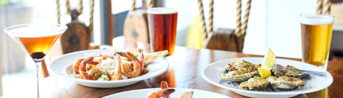 Three plates of seafood dishes and three drinks, including a cocktail and two beers, are set on a wooden table in a bright restaurant with decorative rope accents in the background.