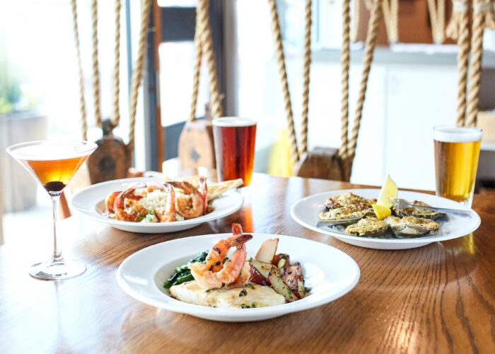 Three plates of seafood dishes and three drinks, including a cocktail and two beers, are set on a wooden table in a bright restaurant with decorative rope accents in the background.