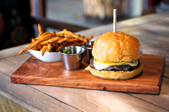A cheeseburger with lettuce and pickles on a brioche bun sits on a wooden board, accompanied by a metal cup of sauce, a bowl of French fries, and sliced pickled jalapeños.