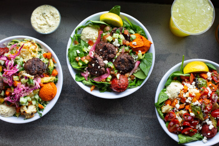 Three bowls of colorful Mediterranean salads with greens, falafel, hummus, vegetables, and lemon wedges, surrounded by a cup of yellow drink and a small bowl of dip, on a gray surface.