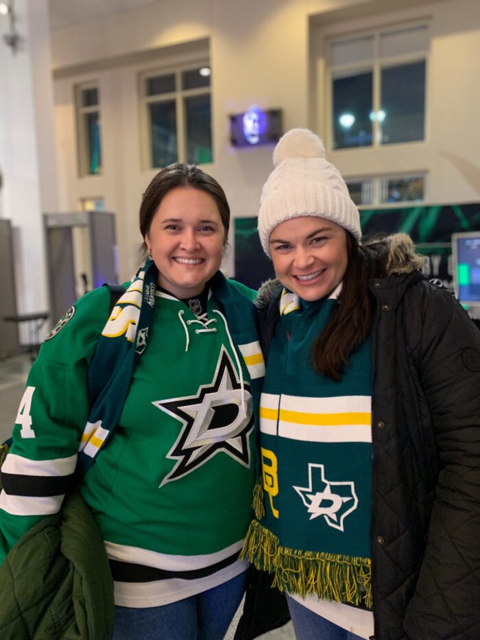Two women smiling, wearing green Dallas Stars hockey jerseys and scarves. One is also wearing a white knit hat and black coat. They are indoors, likely at a hockey arena.