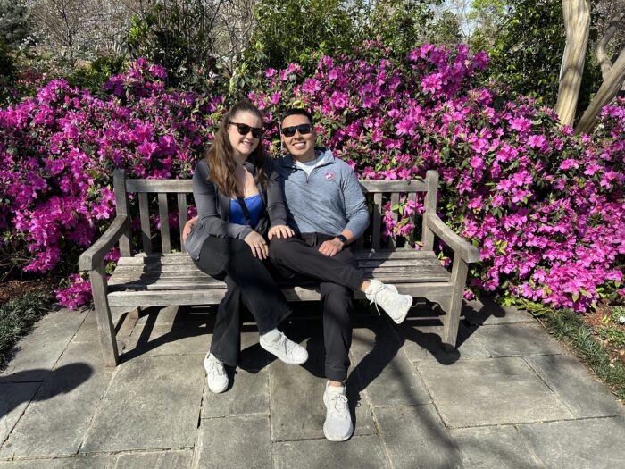 A smiling couple sits on a wooden bench in front of vibrant pink flowers, wearing sunglasses and casual clothes, enjoying a sunny day outdoors.
