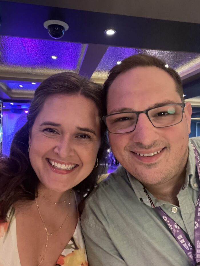 A woman and a man smile closely together for a selfie indoors under purple and yellow lighting; both appear happy and dressed casually.