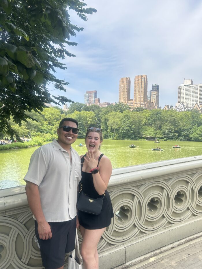 A smiling couple stands on a bridge with a green lake and tall city buildings in the background. The woman holds up her hand to show a ring, suggesting an engagement. Trees and blue sky complete the scenic urban park setting.