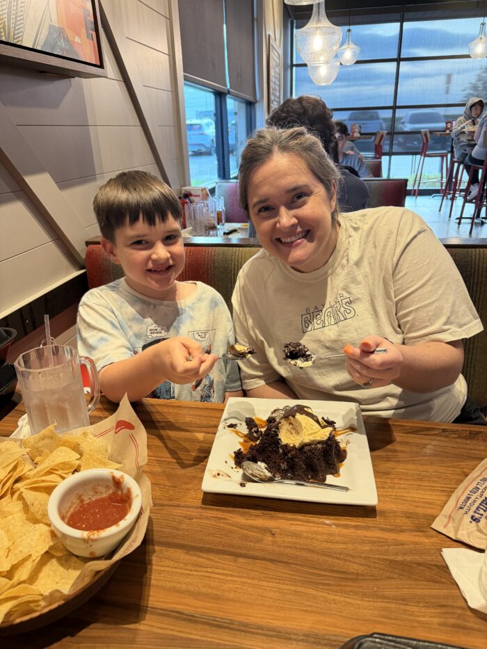 A smiling woman and child sit in a restaurant booth, sharing a dessert with ice cream and chocolate. Chips and salsa are on the table beside them. Both are holding spoons and looking at the camera.