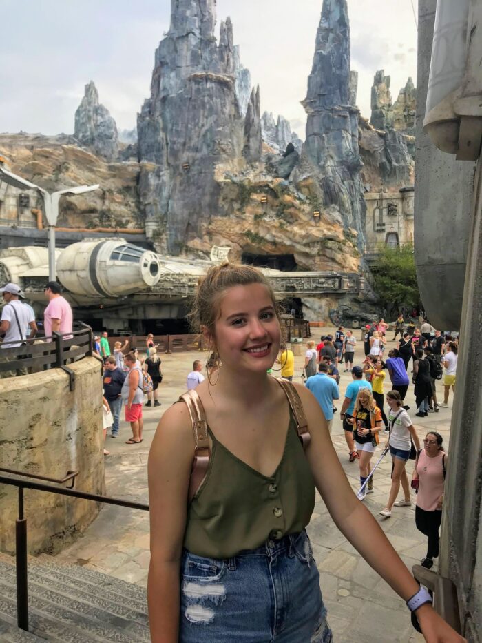 A young woman with a green top and denim shorts smiles at an outdoor Star Wars-themed park with rocky spires, the Millennium Falcon, and many visitors in the background.