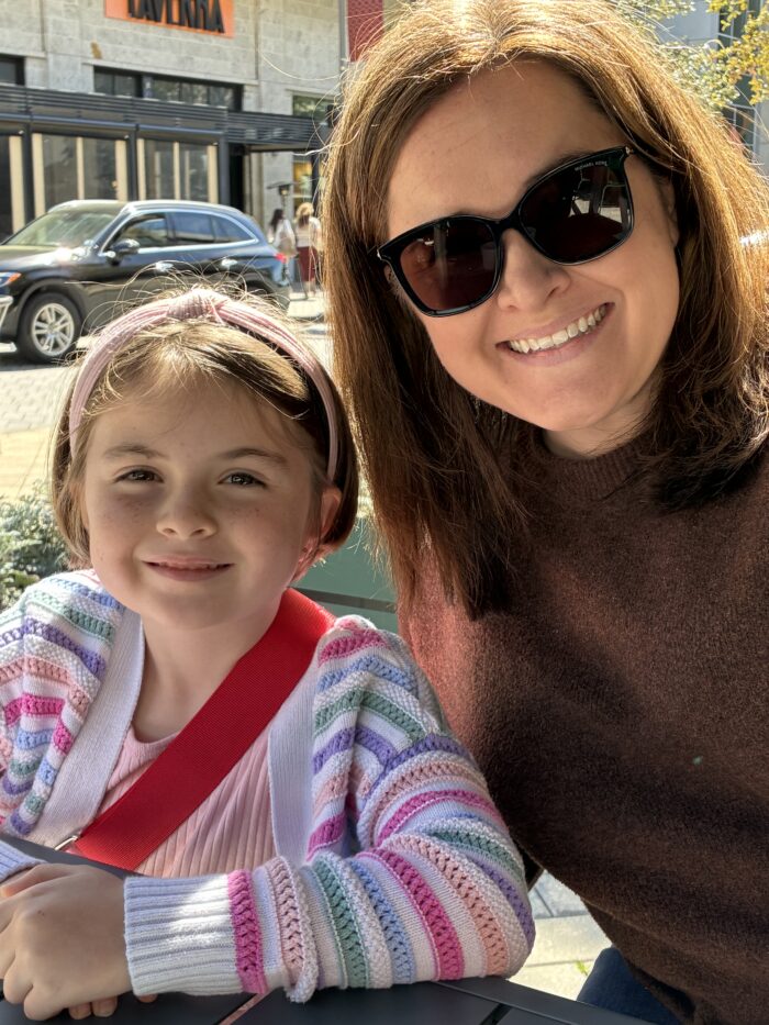 A woman wearing sunglasses and a brown sweater sits next to a young girl in a colorful striped cardigan. They are smiling outdoors at a table, with sunlight shining and cars and buildings in the background.