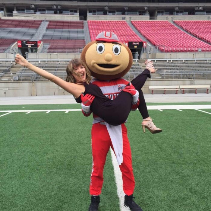 A person in a Brutus Buckeye mascot costume holds a smiling woman in their arms on a football field in an empty stadium. The woman has one arm and one leg extended playfully.