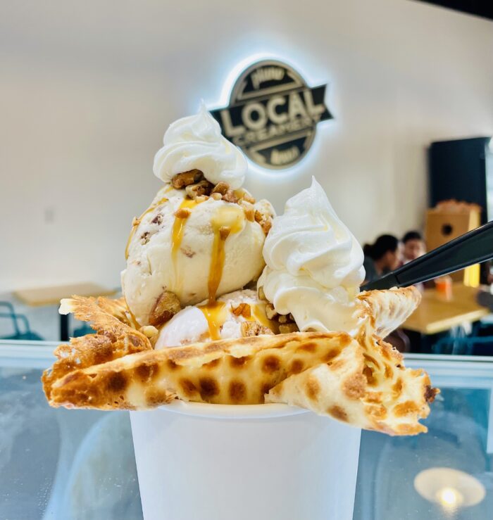 A cup of ice cream topped with whipped cream, caramel sauce, and crumbled nuts, served with waffle pieces. The background features a blurred café setting and a LOCAL sign, highlighting the perfect spot to enjoy your favorite ice cream treats.