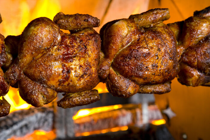 Golden brown rotisserie chickens roasting over an open flame, with crispy skin and visible seasoning, set against a background of glowing orange fire.
