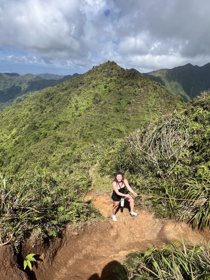 A person in athletic clothing holds a rope while hiking up a steep, dirt trail surrounded by lush green mountains under a partly cloudy sky.