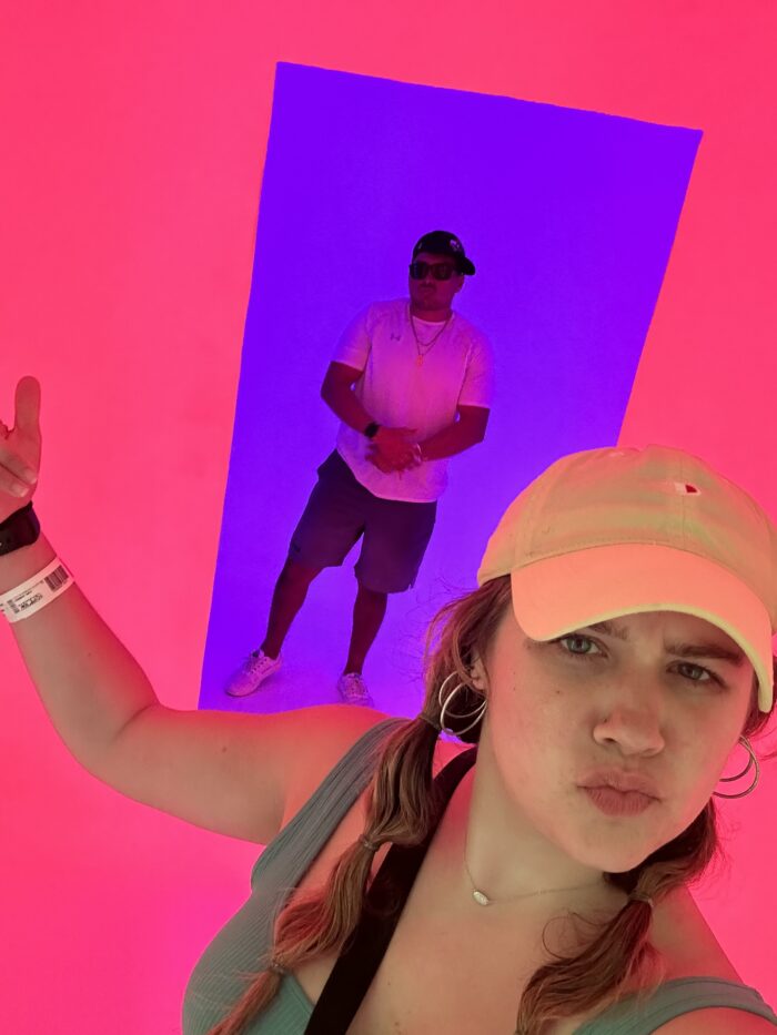A woman in a yellow cap poses for a selfie with a pout, while a man in sunglasses stands in the background. The scene is bathed in bright pink and purple lighting, creating a vibrant, colorful atmosphere.
