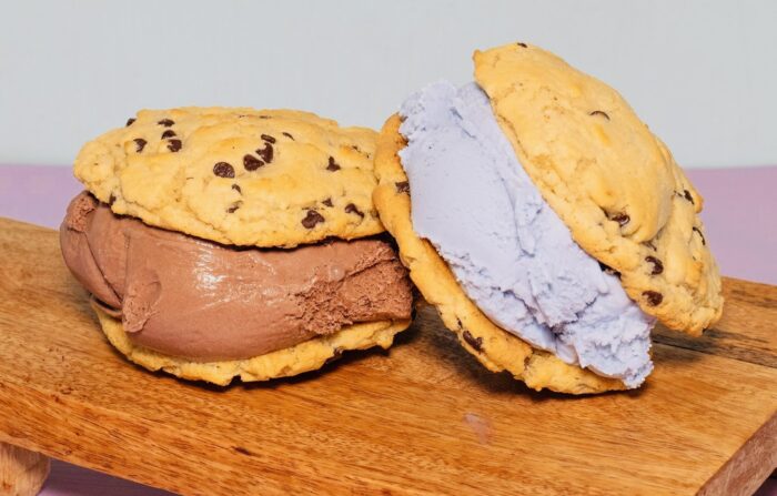 Two ice cream sandwiches made with chocolate chip cookies—one filled with chocolate ice cream, the other with purple ice cream—are placed on a wooden board against a light background, showcasing the perfect ice cream treat.