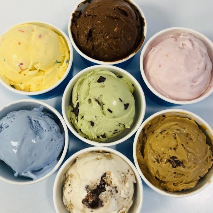 Seven cups of colorful Ice Cream are arranged in a circle. Flavors appear to include chocolate, strawberry, mint chip, salted caramel, funfetti, blue, and cookies and cream. The background is a light, plain surface.