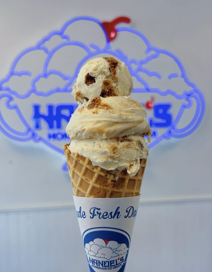 A close-up of a waffle cone filled with two scoops of ice cream, held in front of a blurred Handels Homemade Ice Cream sign. A cherry sits on top of the ice cream, and the cone is wrapped in branded paper.