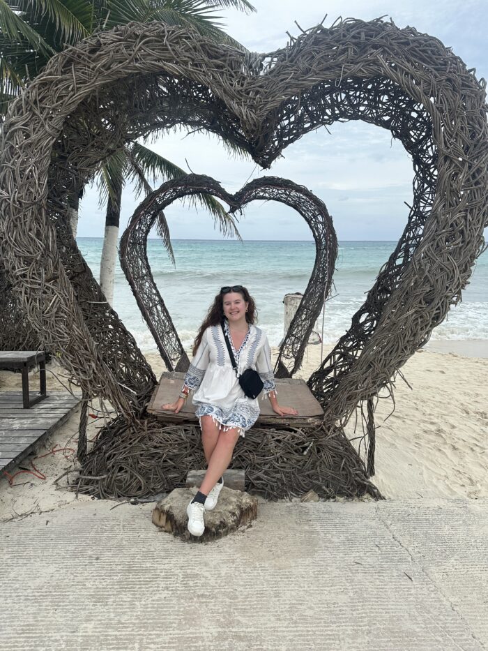 A woman in a white dress sits smiling on a swing framed by two large heart-shaped arches made of intertwined branches, with a beach, palm tree, and ocean in the background.