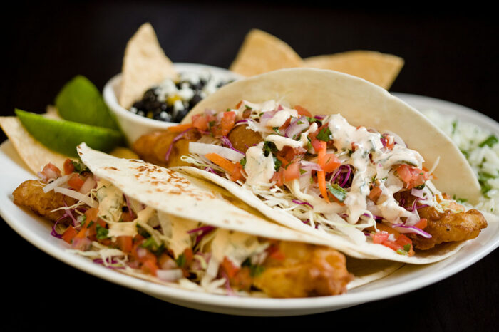 Two fish tacos topped with shredded cabbage, diced tomatoes, and creamy sauce are served on a white plate with lime wedges, tortilla chips, and a small bowl of black beans in the background.