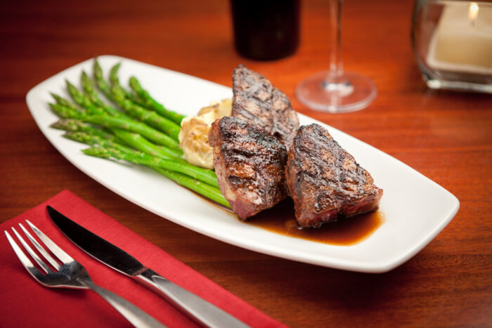 Grilled steak with grill marks served on a white plate with asparagus spears, mashed potatoes, and sauce, set on a wooden table with a fork, knife, red napkin, wine glass, and candle in the background.