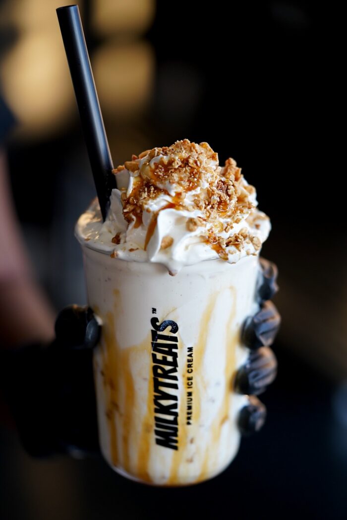 A gloved hand holds a MilkyTreats cup filled with a creamy milkshake topped with whipped cream, caramel drizzle, and crunchy nut pieces, with a black straw inserted.