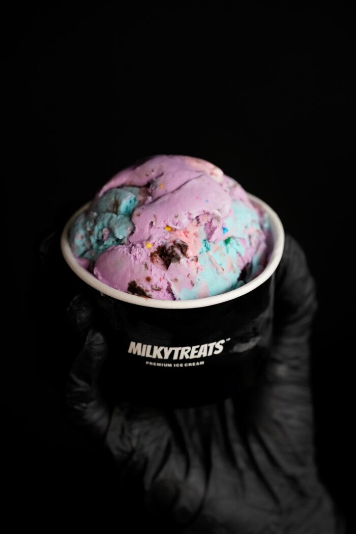 A black-gloved hand holds a black cup labeled MILKYTREATS containing a scoop of colorful ice cream in shades of purple, blue, and pink against a black background.