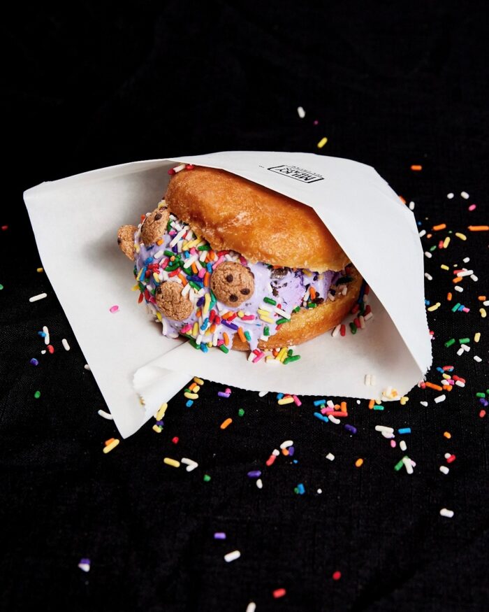A donut sliced in half and filled with purple ice cream, rainbow sprinkles, and mini cookies, partially wrapped in white paper, with sprinkles scattered on a black surface.