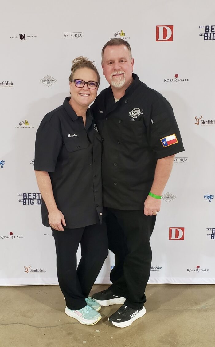 A smiling man and woman stand arm-in-arm on a red carpet in black chef jackets, the man sporting a Texas flag patch. A step-and-repeat banner with various logos is behind them, hinting at an event celebrating food like ice cream.
