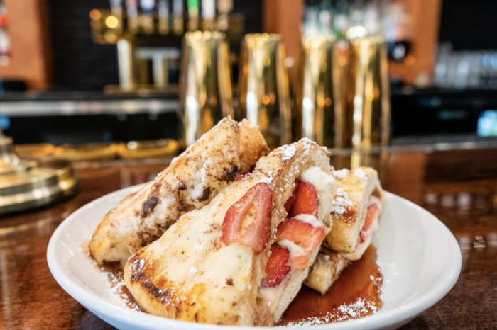 A plate of French toast stuffed with cream and fresh strawberries, topped with powdered sugar and served with syrup, sits on a wooden bar with golden drink shakers in the background.