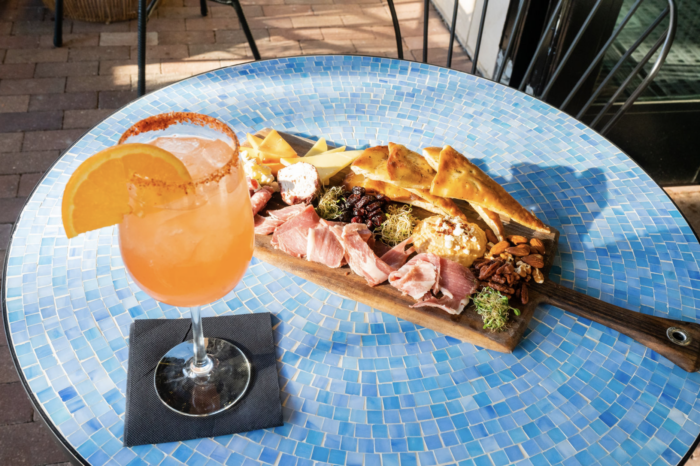 A cocktail garnished with an orange slice sits on a blue mosaic table next to a wooden charcuterie board with meats, cheeses, crackers, nuts, and fruit.
