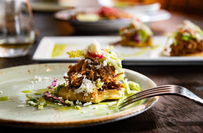 A close-up of a plated dish with shredded meat, lettuce, radish, and crumbled cheese on a round corn cake, with a fork on the side. Similar dishes are blurred in the background.