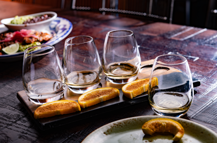 Four glasses of tequila, some partially filled, are arranged on a wooden tray with orange slices. A plate with food is blurred in the background, and another orange slice sits on a plate in the foreground.