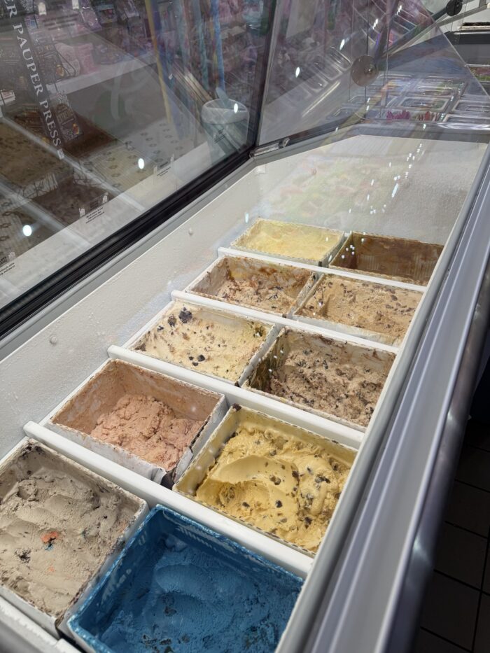 An ice cream display case filled with various ice cream flavors in rectangular tubs, including chocolate chip, cookie dough, mint, and more, inside a brightly lit ice cream shop.