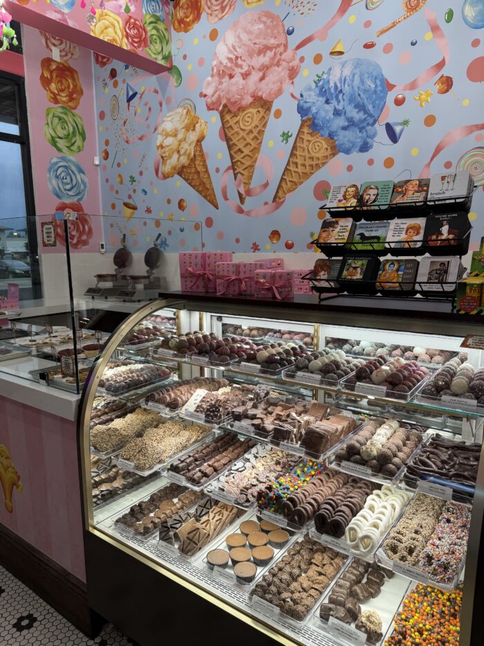 A brightly decorated candy shop displays a glass case filled with assorted chocolates and sweets. The wall behind features colorful ice cream cone art and festive patterns, while shelves with boxed treats and ice cream delights sit above the case.
