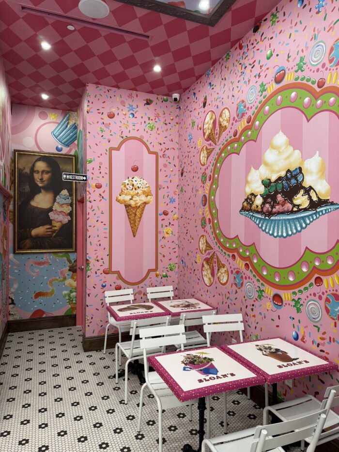 A colorful ice cream-themed café with pink walls decorated with ice cream art, sprinkles, and a playful Mona Lisa portrait holding an ice cream cone. White tables and chairs sit on a black-and-white tiled floor, creating the perfect spot to enjoy your favorite ice cream.