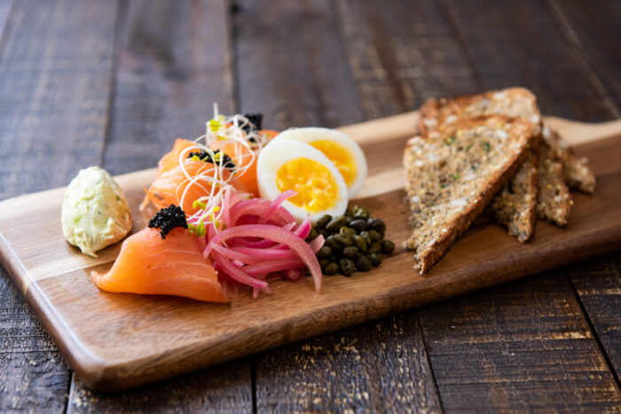 A wooden board with smoked salmon, sliced boiled eggs, pickled red onions, capers, sprouts, black caviar, herbed cream cheese, and toasted multigrain bread on a rustic wooden table.