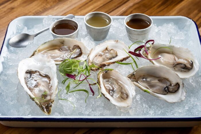 A tray with six raw oysters on the half shell served on crushed ice, garnished with microgreens, accompanied by three metal cups of sauces and a small spoon, on a wooden table.