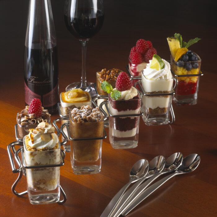 A selection of colorful layered mini desserts in shot glasses, arranged on a wooden table with silver spoons, a bottle of red wine, and a filled wine glass in the background. Each dessert is topped with fruit or cream.
