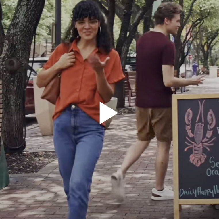 A woman in an orange shirt and blue jeans walks on a brick sidewalk, smiling and gesturing as if celebrating a soccer goal. Behind her, a man in a maroon shirt and shorts passes a chalkboard sign with a drawing of a lobster. Trees and parked cars are visible.