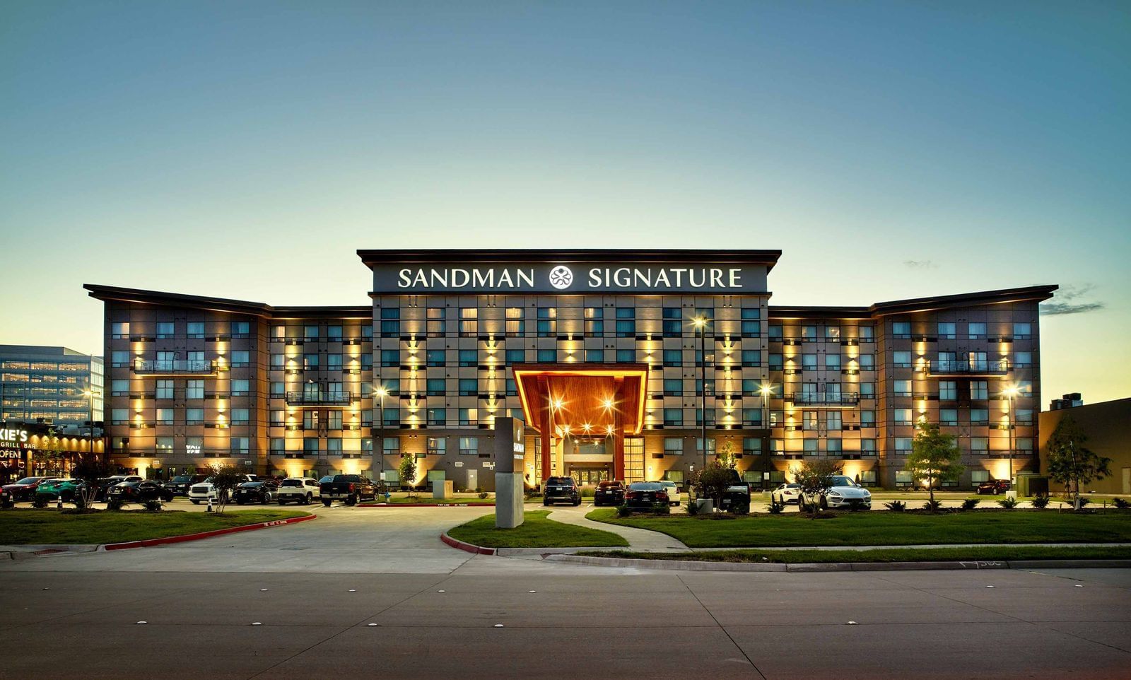 Modern, well-lit hotel building at dusk with Sandman Signature sign on top, large windows, illuminated entrance canopy, and cars parked in front on a curved driveway—perfect for travelers seeking comfort and exclusive hotel deals.