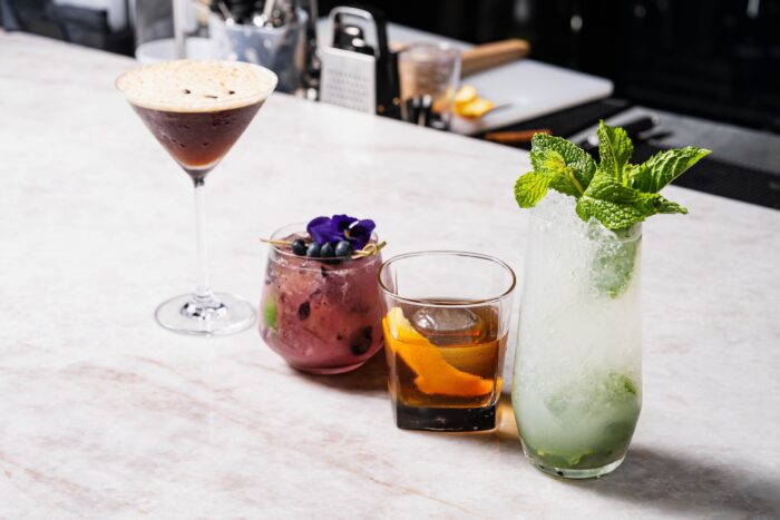 Four cocktails, including an espresso martini, a berry drink with a flower, an old fashioned with an olive and fig garnish, and a mint-topped mojito, arranged on a marble bar top.
