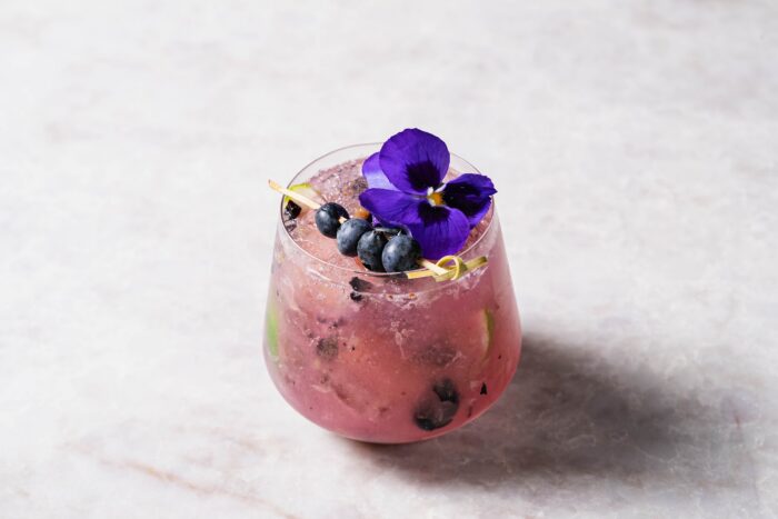 A pink Olive and Fig cocktail in a glass garnished with blueberries and a purple edible flower.